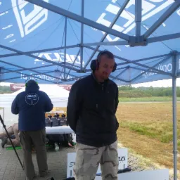 Strzelnica: mężczyzna w słuchawkach stoi pod namiotem, w tle osoby i karabiny na stole. Widok na poligon i zalesiony teren.