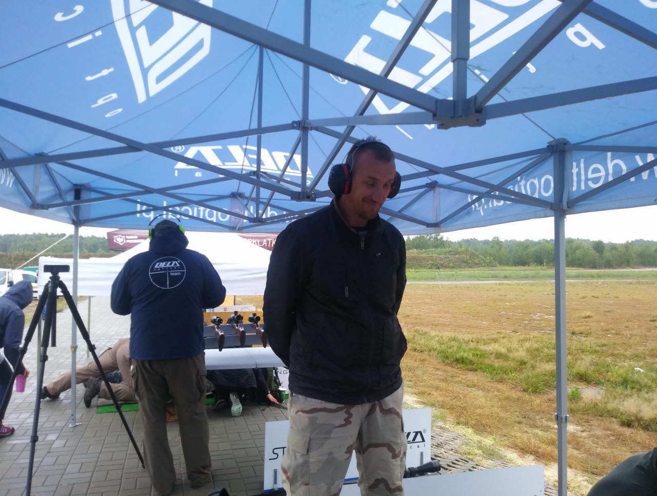 Strzelnica: mężczyzna w słuchawkach stoi pod namiotem, w tle osoby i karabiny na stole. Widok na poligon i zalesiony teren.