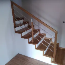 Nowoczesne schody z drewnianymi stopniami i białymi podstopnicami, zabezpieczone metalową balustradą z drewnianym pochwytem, widoczne z dołu pod kątem w jasnym wnętrzu.