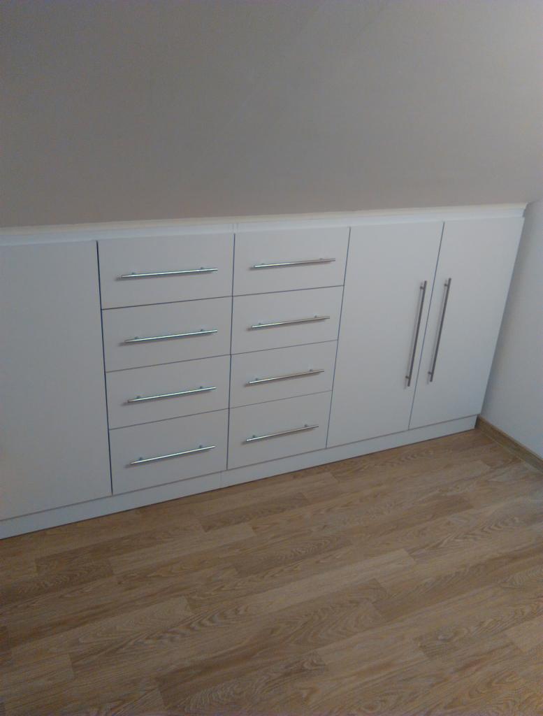 Biała zabudowa meblowa ze srebrnymi uchwytami, wkomponowana w skos poddasza, z drewnianą podłogą.