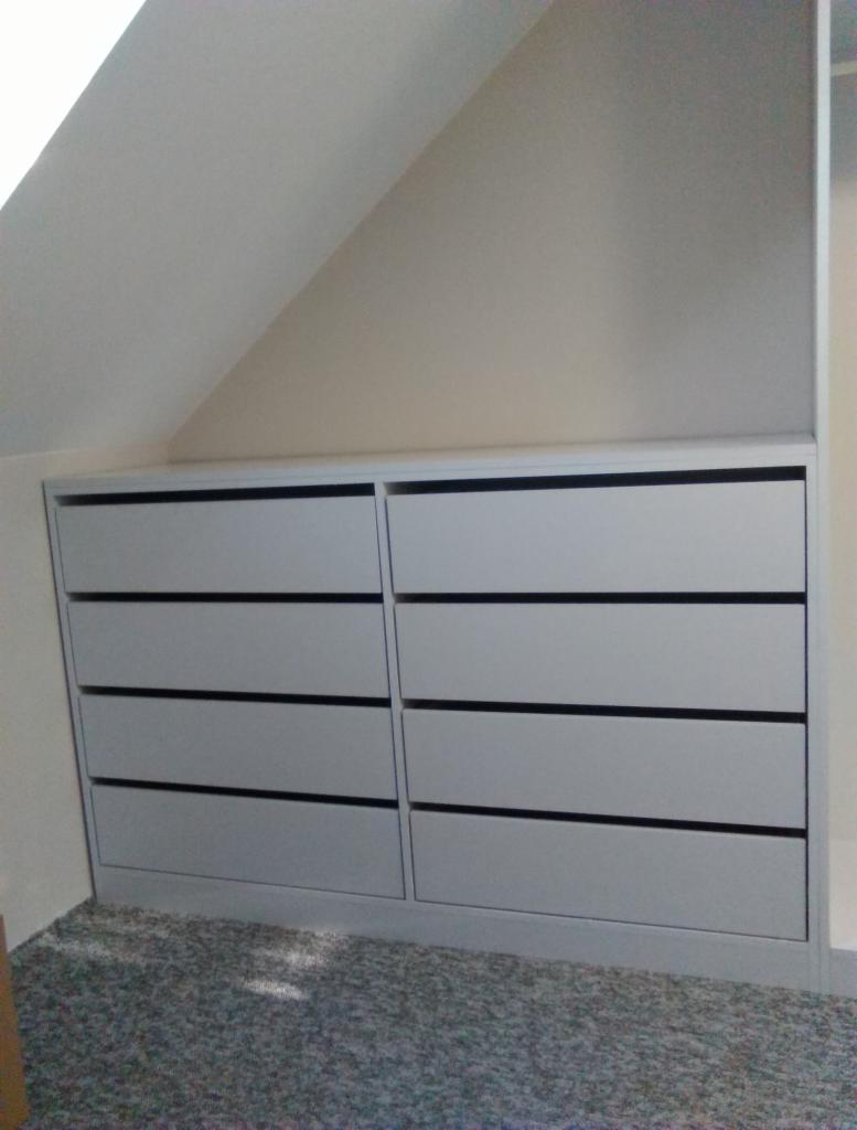 Szara zabudowa pod skosem z czterema szufladami z czarnymi wpuszczonymi uchwytami, dopasowana do przestrzeni poddasza.