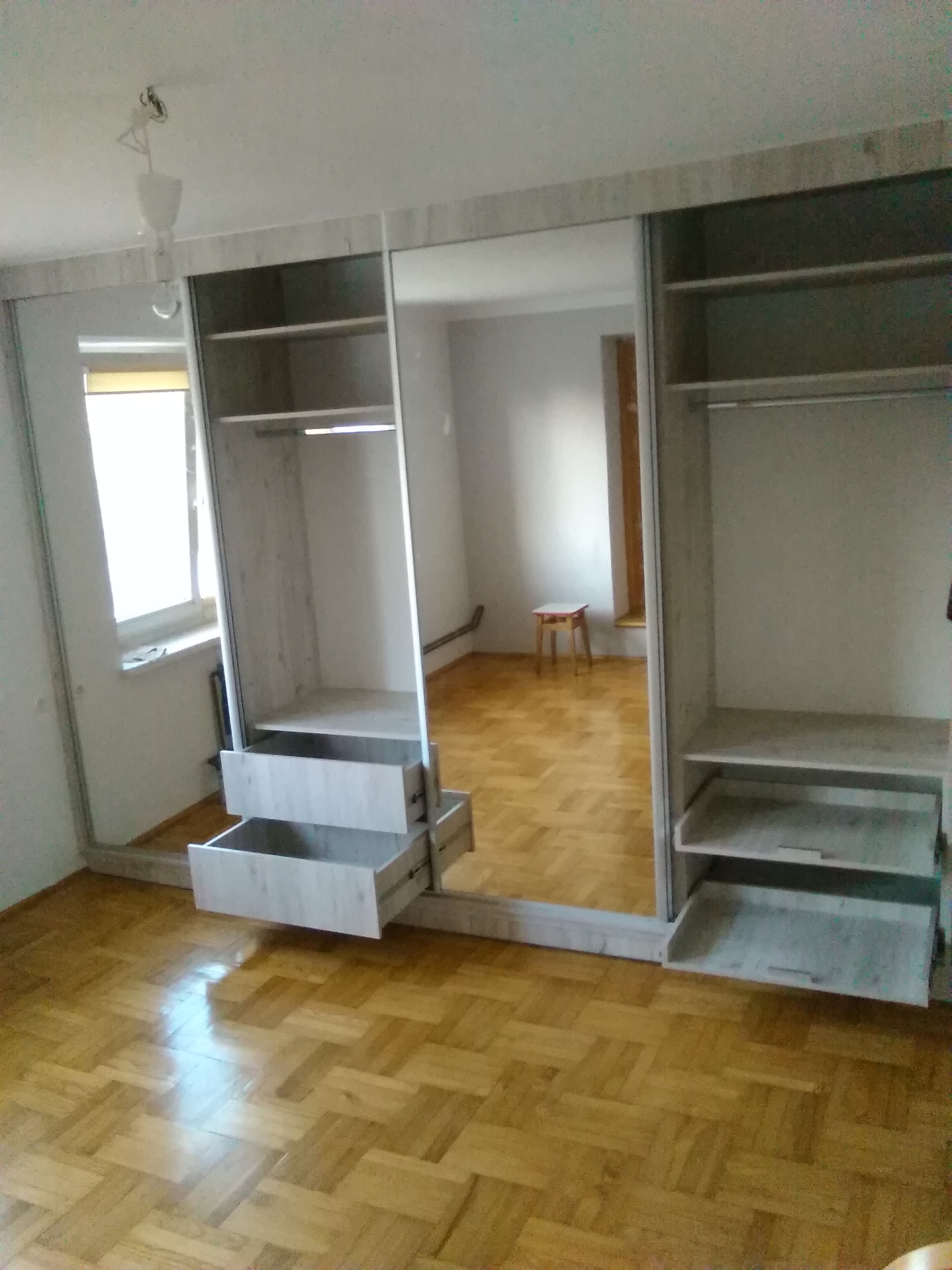 Szafa wnękowa z przesuwnymi drzwiami lustrzanymi i wysuwanymi szufladami w odcieniach szarości, na tle parkietu, z widocznym odbiciem pokoju z krzesłem.