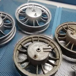 Cztery felgi motocyklowe, dwie po lewej stronie w kolorze srebrnym, dwie po prawej w stanie surowym, leżące na perforowanym stole warsztatowym z widocznym pistoletem do piaskowania.