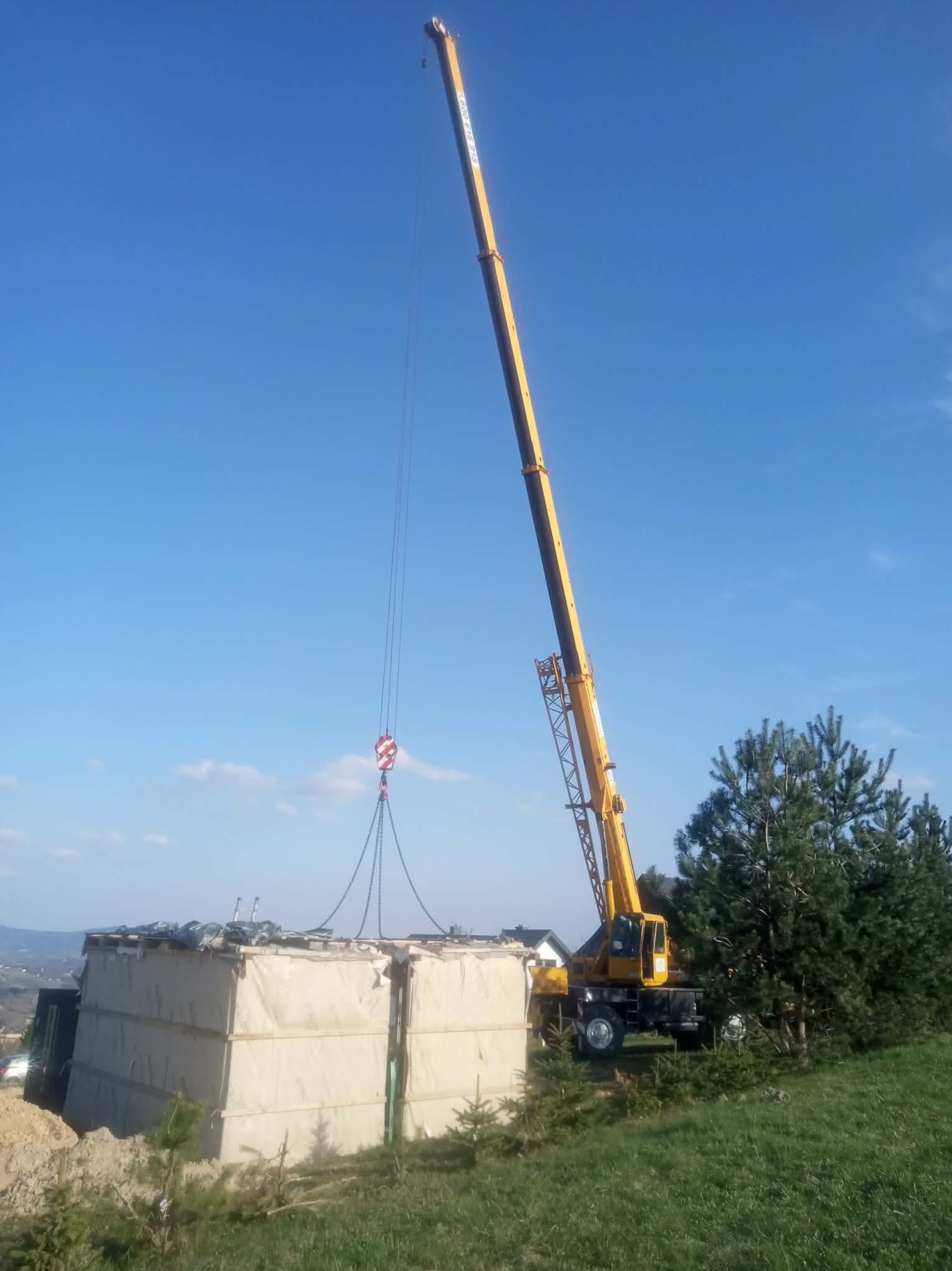 Żółty dźwig podnosi duży, jasny element na tle błękitnego nieba i zieleni. Widoczne drzewa i oddalony krajobraz. Praca w toku, etap transportu elementu.