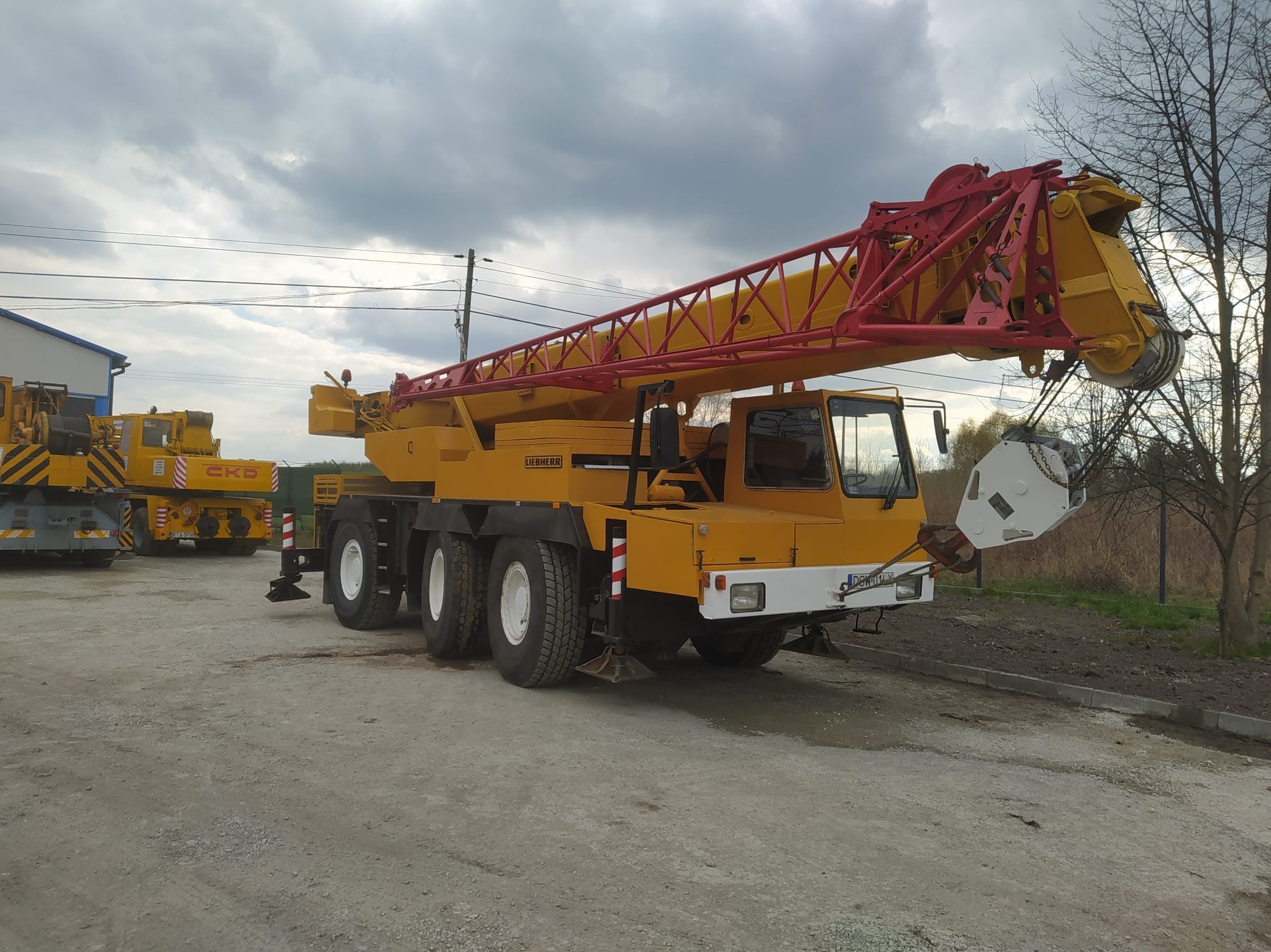 Żółto-czerwony dźwig Liebherr na placu w Wojniczu, widoczny z boku, z wysuniętym ramieniem. W tle drugi dźwig i pochmurne niebo.