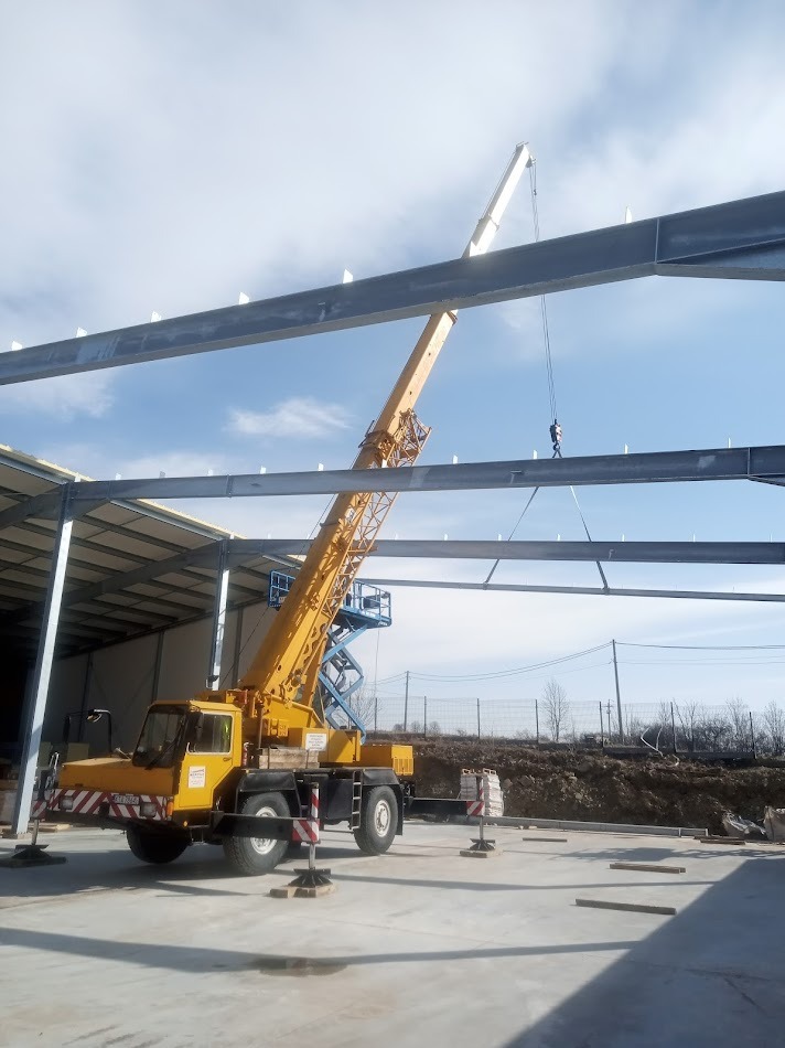 Żółty dźwig samochodowy z wysięgnikiem podnosi stalową konstrukcję hali. W tle błękitne niebo z chmurami i zarys drzew. Widoczny podnośnik koszowy.