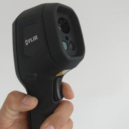 Paweł Drewniak - Czarny termowizor marki FLIR trzymany w dłoni na tle jasnej ściany.