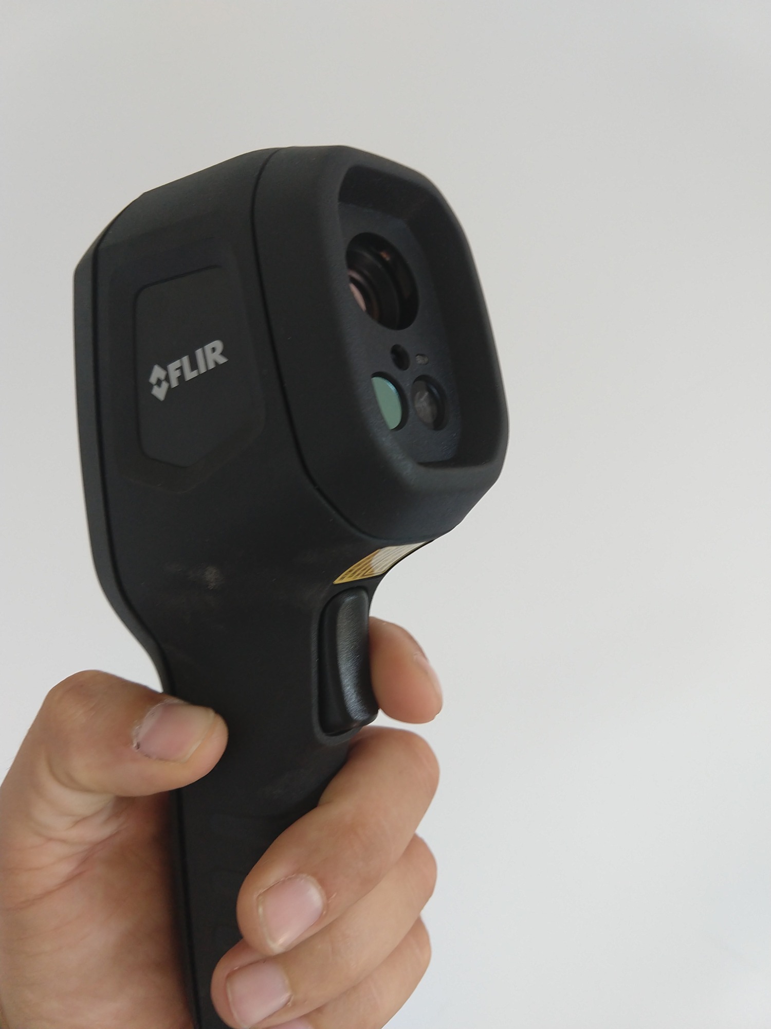 Czarny termowizor marki FLIR trzymany w dłoni na tle jasnej ściany.