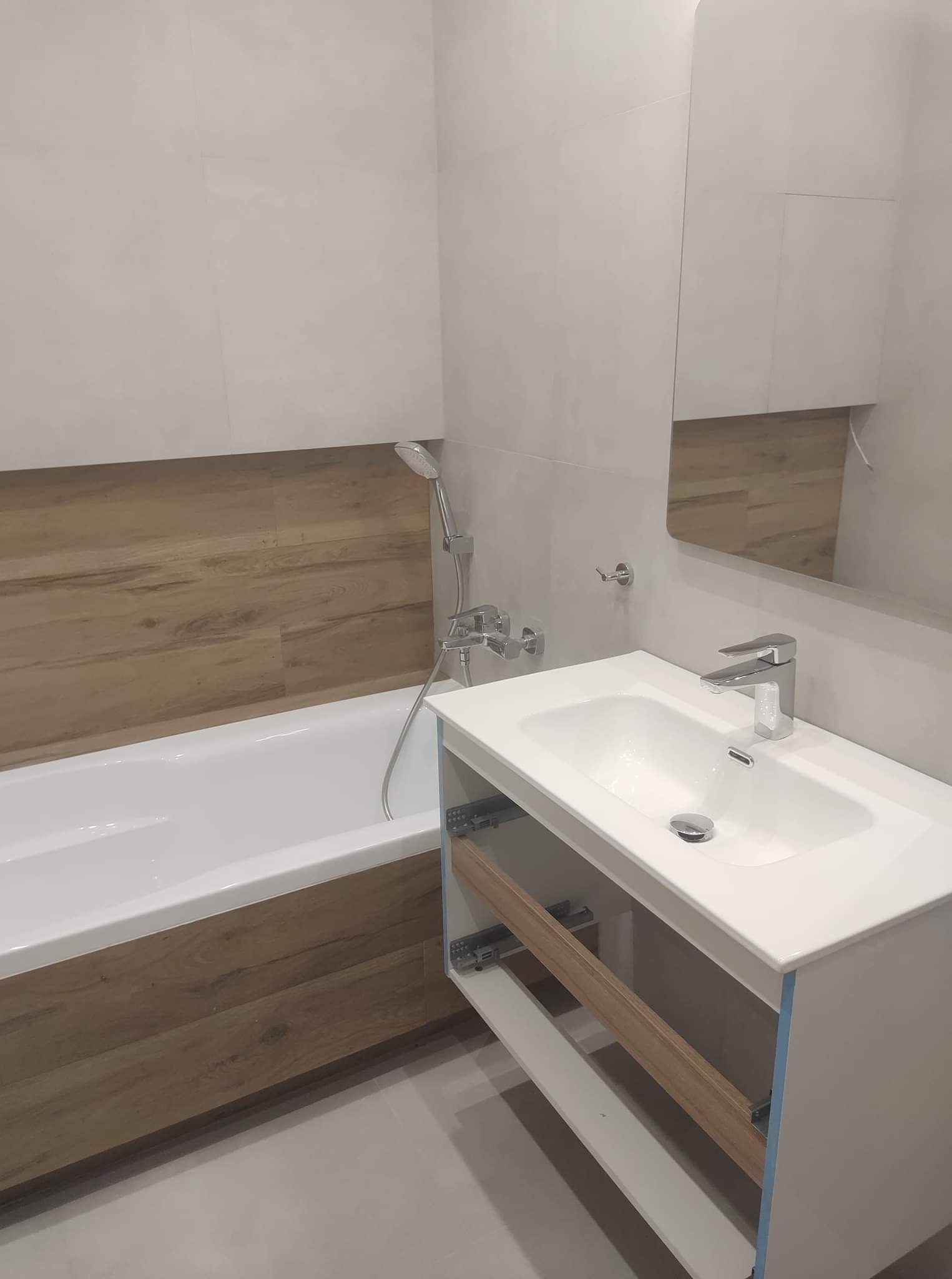 Nowoczesna łazienka z wanną obudowaną panelami imitującymi drewno, białą umywalką z szafką bez frontów szuflad i lustrem na ścianie z jasnymi płytkami.