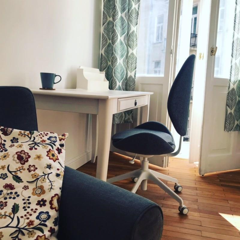 Gabinet z biurkiem, krzesłem ergonomicznym i sofą z poduszką w kwiaty, widok na balkon przez otwarte drzwi.