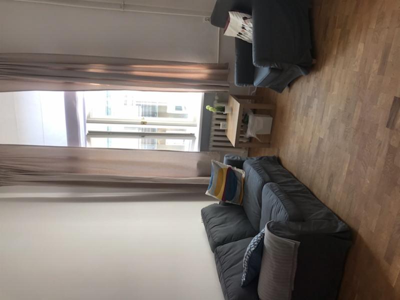 Pokój dzienny z szarą sofą, fotelami i drewnianą podłogą, oświetlony naturalnym światłem z okna z zasłonami.