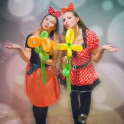 Dwie uśmiechnięte kobiety w kostiumach w stylu Myszki Miki pozują z balonowymi kwiatami, otoczone unoszącymi się bańkami mydlanymi na rozmytym tle.