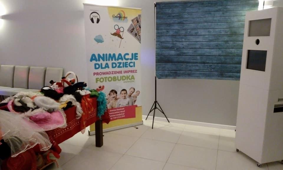 Stanowisko z fotobudką, rekwizytami na stole przykrytym czerwoną tkaniną i banerem reklamowym z napisem 'Animacje dla dzieci. Prowadzenie imprez. Fotobudka Nowość'