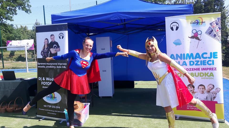 Dwie uśmiechnięte kobiety w kostiumach superbohaterów, na tle stoiska z banerami reklamującymi oprawę muzyczną imprez, animacje dla dzieci i fotobudkę.