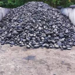 Góra antracytu w betonowym boksie z widocznymi oponami samochodowymi na górnej krawędzi ściany, pod drzewami.