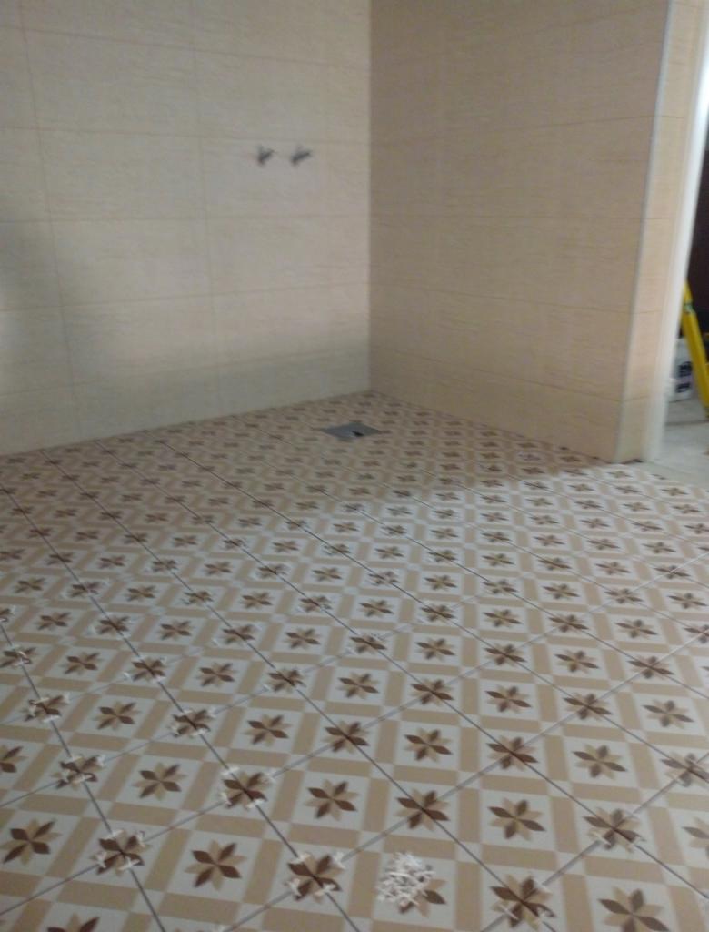 Wykończona łazienka z beżowymi płytkami podłogowymi w kwiaty i jasnymi płytkami ściennymi, widoczne dwa uchwyty na ścianie i odpływ podłogowy.