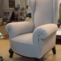 Odnowiony fotel typu uszak w jasnej tapicerce z wyraźnym splotem, widoczne niedoskonałości na podłokietniku, stojący w warsztacie tapicerskim z narzędziami w tle.