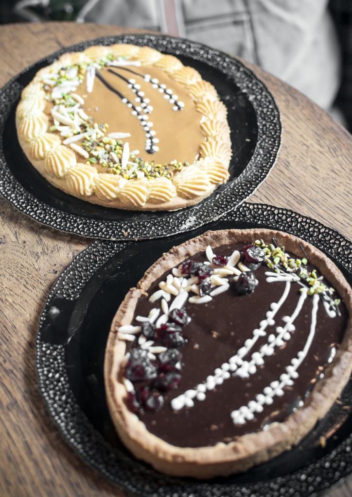 Dwa okrągłe tarty na czarnych dekoracyjnych talerzach: karmelowa z kremowymi rozetkami, migdałami i pistacjami oraz czekoladowa z wiśniami, migdałami i pistacjami, obie ozdobione białymi kropkami.