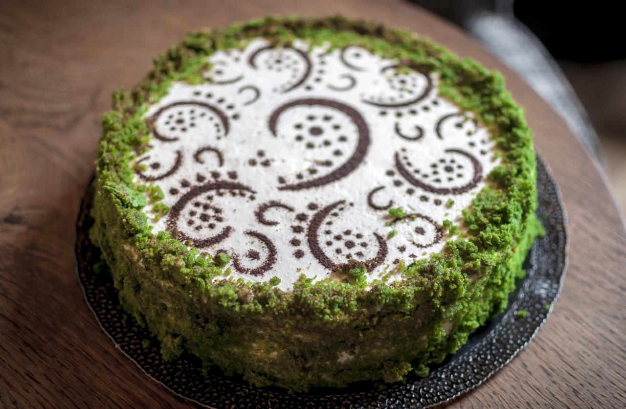 Okrągły tort z białym kremem i zieloną posypką, dekorowany ciemnymi wzorami w kształcie zawijasów i kropek.