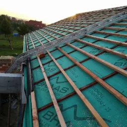 Konstrukcja dachu z zieloną membraną izolacyjną, drewnianymi łatami i aluminiową drabinką dekarską w trakcie budowy, widok z g&oacute;ry na tle zachodzącego słońca.