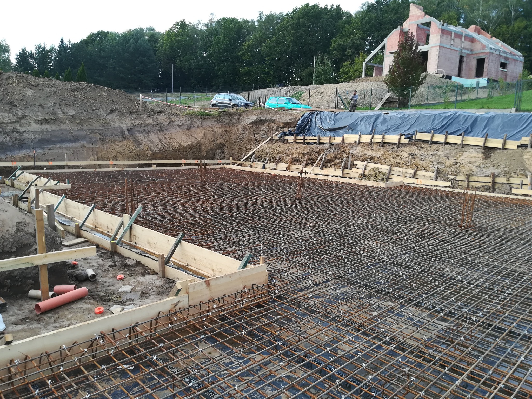 Przygotowanie zbrojenia i szalunku pod fundamenty domu jednorodzinnego na działce z widoczną konstrukcją budynku w tle.