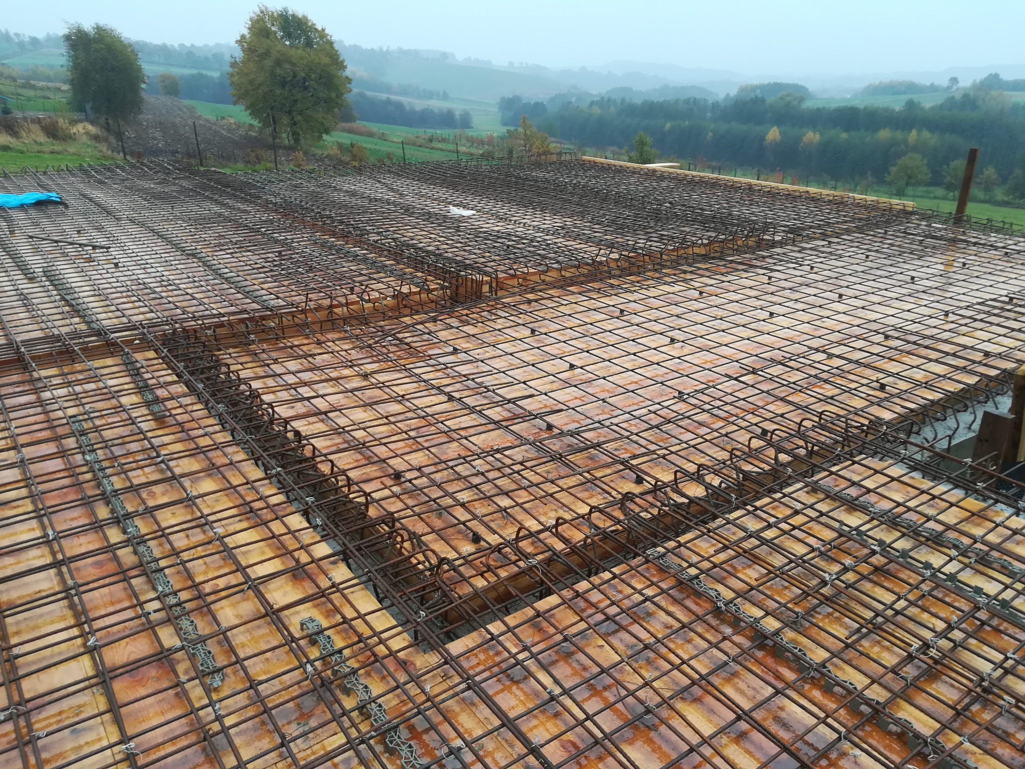 Zbrojenie stropu na deskowaniu z widocznymi dystansami i otaczającym krajobrazem w tle w pochmurny dzień.