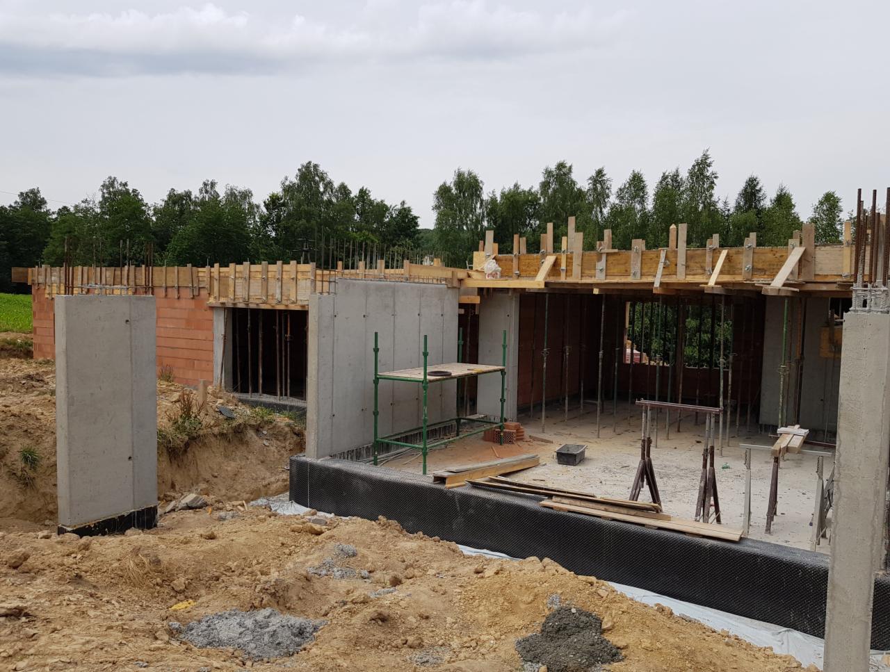 Surowy stan budowy domu jednorodzinnego: betonowe ściany fundamentowe z izolacją przeciwwilgociową, szalunki stropowe na podporach, częściowo wymurowane ściany z czerwonej cegły.