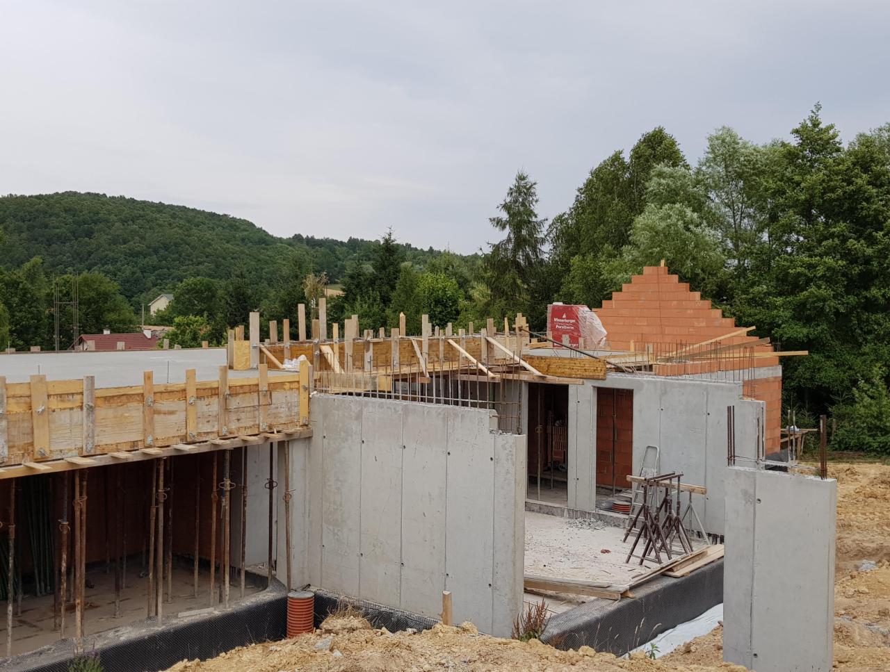Budowa domu jednorodzinnego w stanie surowym otwartym: betonowe ściany fundamentowe, szalunki stropu, mur z cegły ceramicznej, widok na zalesione wzgórze w tle.
