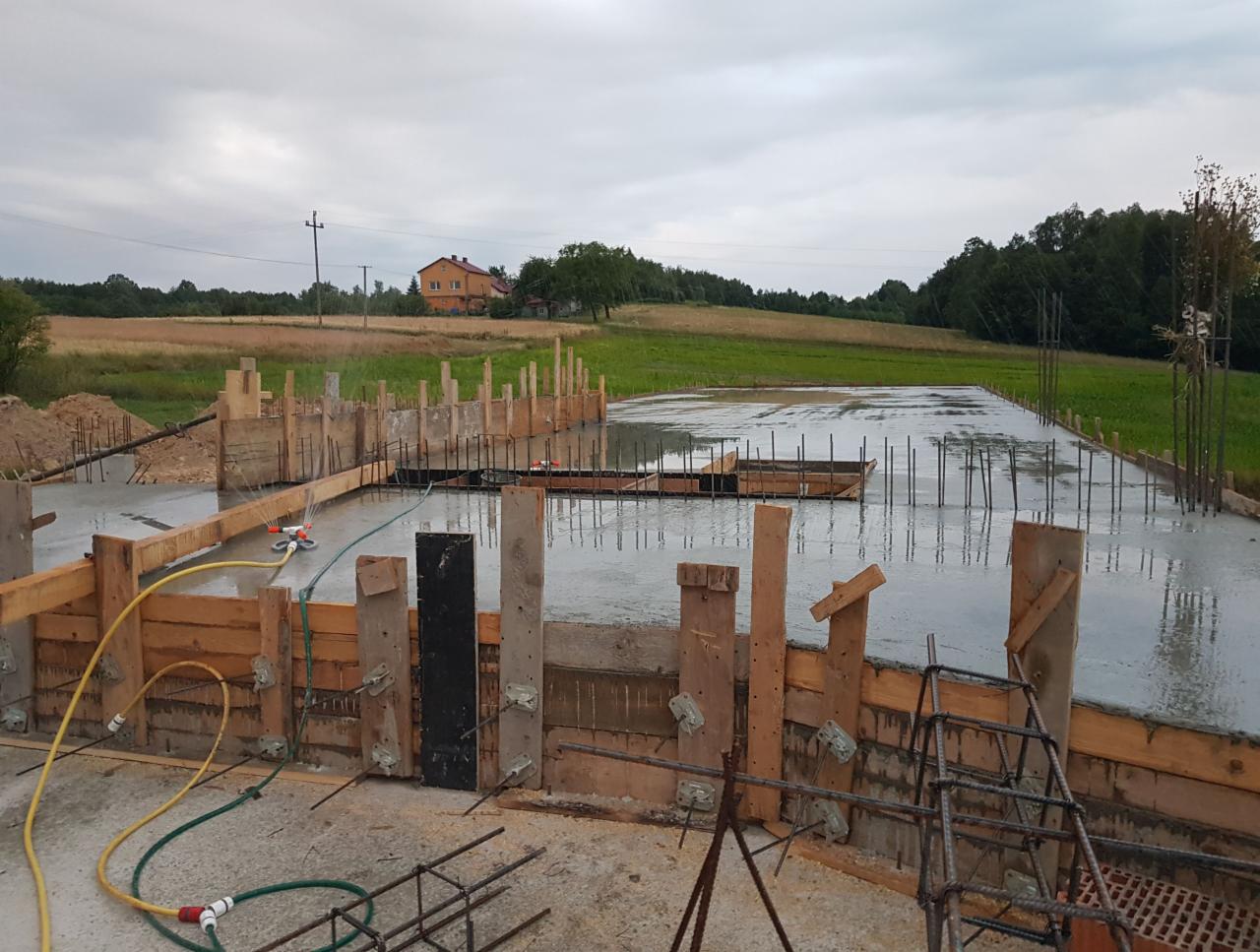 Wylany fundament z drewnianym szalunkiem, widoczne zbrojenia i węże do podlewania betonu, w tle krajobraz wiejski z domem.