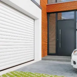 Nowoczesny dom jednorodzinny z białą segmentową bramą garażową, grafitowymi drzwiami wejściowymi z długim uchwytem i drewnianą elewacją, częściowo widoczny biały samochód.