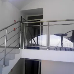 Wewnętrzna klatka schodowa z balustradą ze stali nierdzewnej i szkła, widoczne schody z metalowymi poręczami i krzesło z małym stolikiem za szklanym panelem.