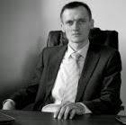 Portret biznesowy mężczyzny w garniturze, siedzącego za biurkiem, z poważnym wyrazem twarzy, w czarno-białej tonacji.