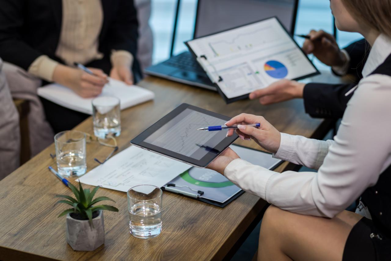 Zespół analizuje dane na tablecie i wykresy podczas spotkania biznesowego przy drewnianym stole, widoczne szklanki z wodą i roślina doniczkowa.