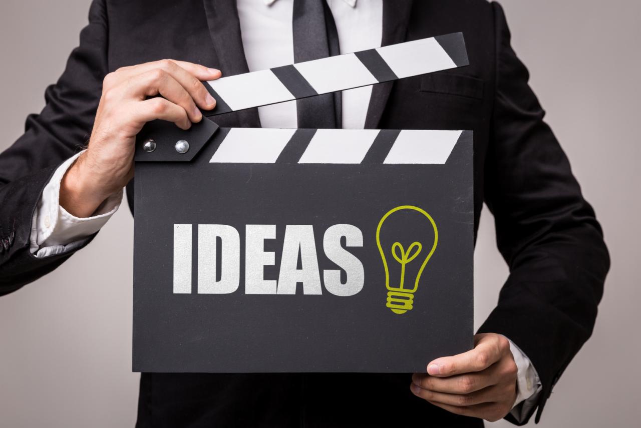 Mężczyzna w garniturze trzyma klaps filmowy z napisem 'IDEAS' i ikoną żarówki, symbolizujący kreatywne pomysły.