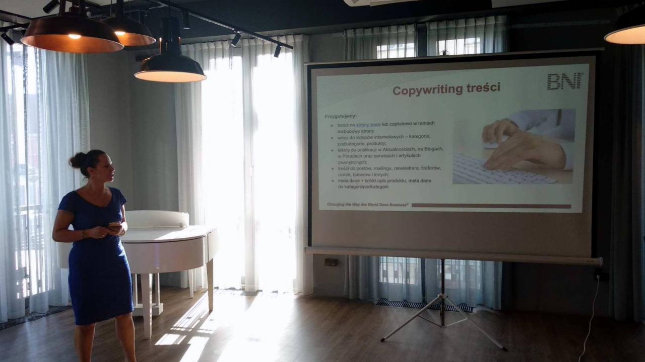 Prezentacja na ekranie projektora dotycząca copywritingu treści, kobieta w niebieskiej sukience stoi obok, w tle designerskie lampy i jasne zasłony.