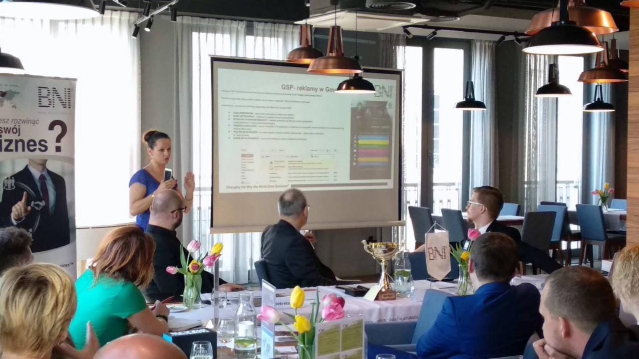 Prezentacja multimedialna o reklamie internetowej podczas spotkania biznesowego, z widocznym ekranem projektora, uczestnikami przy stołach i dekoracjami kwiatowymi.