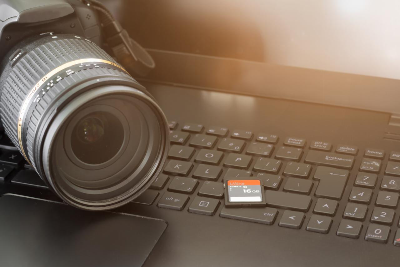Aparat DSLR z obiektywem Tamron leżący na klawiaturze laptopa, obok karta pamięci SD 16GB. Sprzęt fotografa.