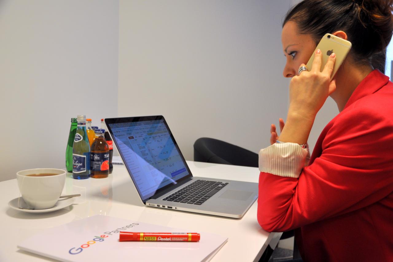 Kobieta w czerwonej marynarce rozmawia przez złoty telefon, na biurku laptop z otwartą stroną internetową, napój, kawa i dokument z logo Google Partners.