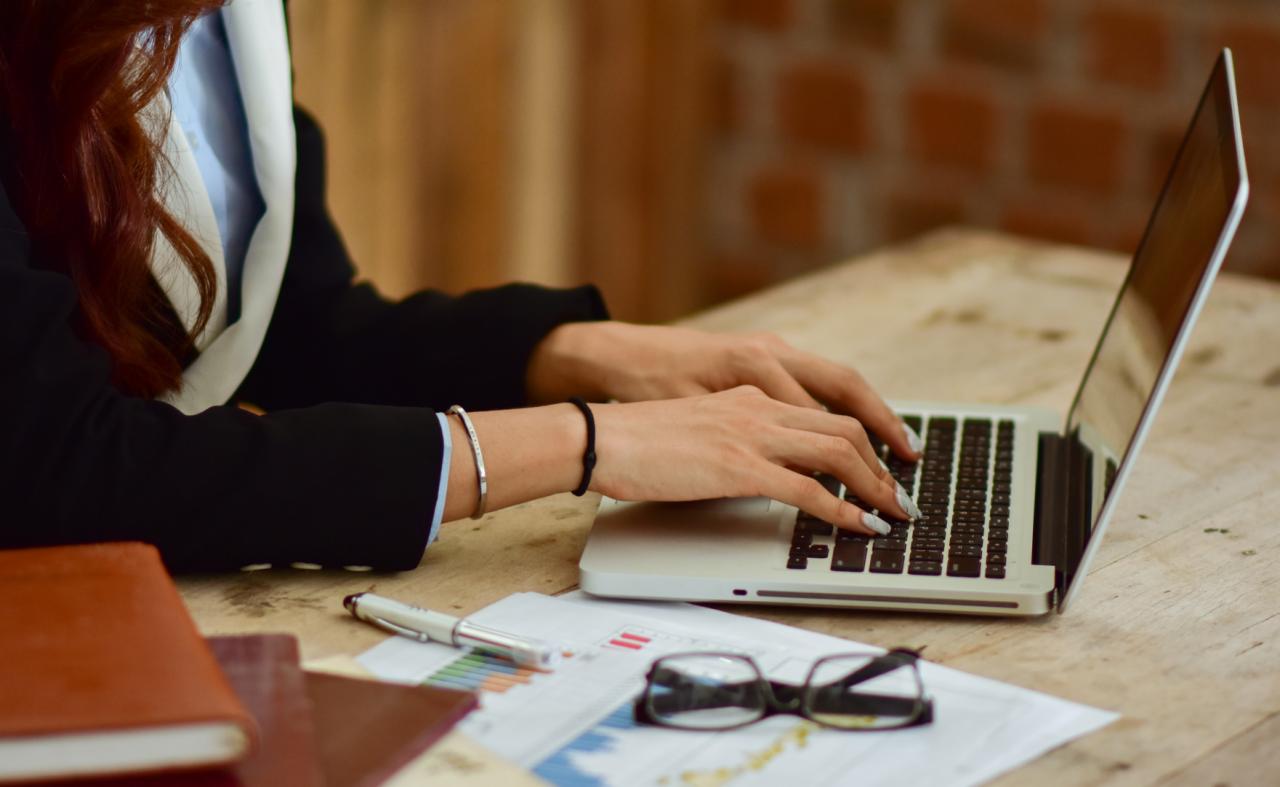 Kobieta w czarnej marynarce pisze na laptopie; widoczne dłonie, fragment laptopa, notatki z wykresami, okulary, długopis i brązowy notes na drewnianym biurku.