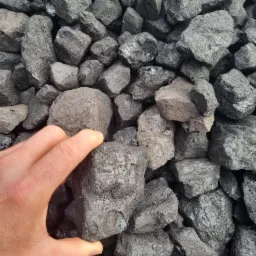 Dłoń trzyma bryłę antracytu na tle sterty węgla, widoczne naturalne pęknięcia i matowa powierzchnia.