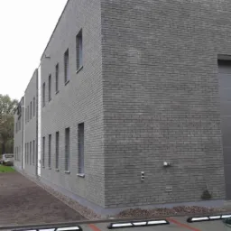 Nowoczesny budynek biurowy z szarej cegły klinkierowej, z widoczną elewacją, oknami i parkingiem z wyznaczonymi miejscami.