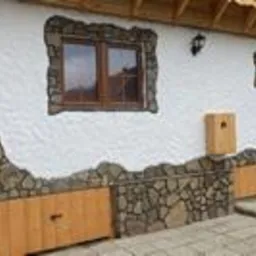 Biała ściana budynku z dekoracyjnym obłożeniem z kamienia i drewnianymi drzwiczkami w dolnej części. Okno z brązową ramą. Nierówna faktura tynku.