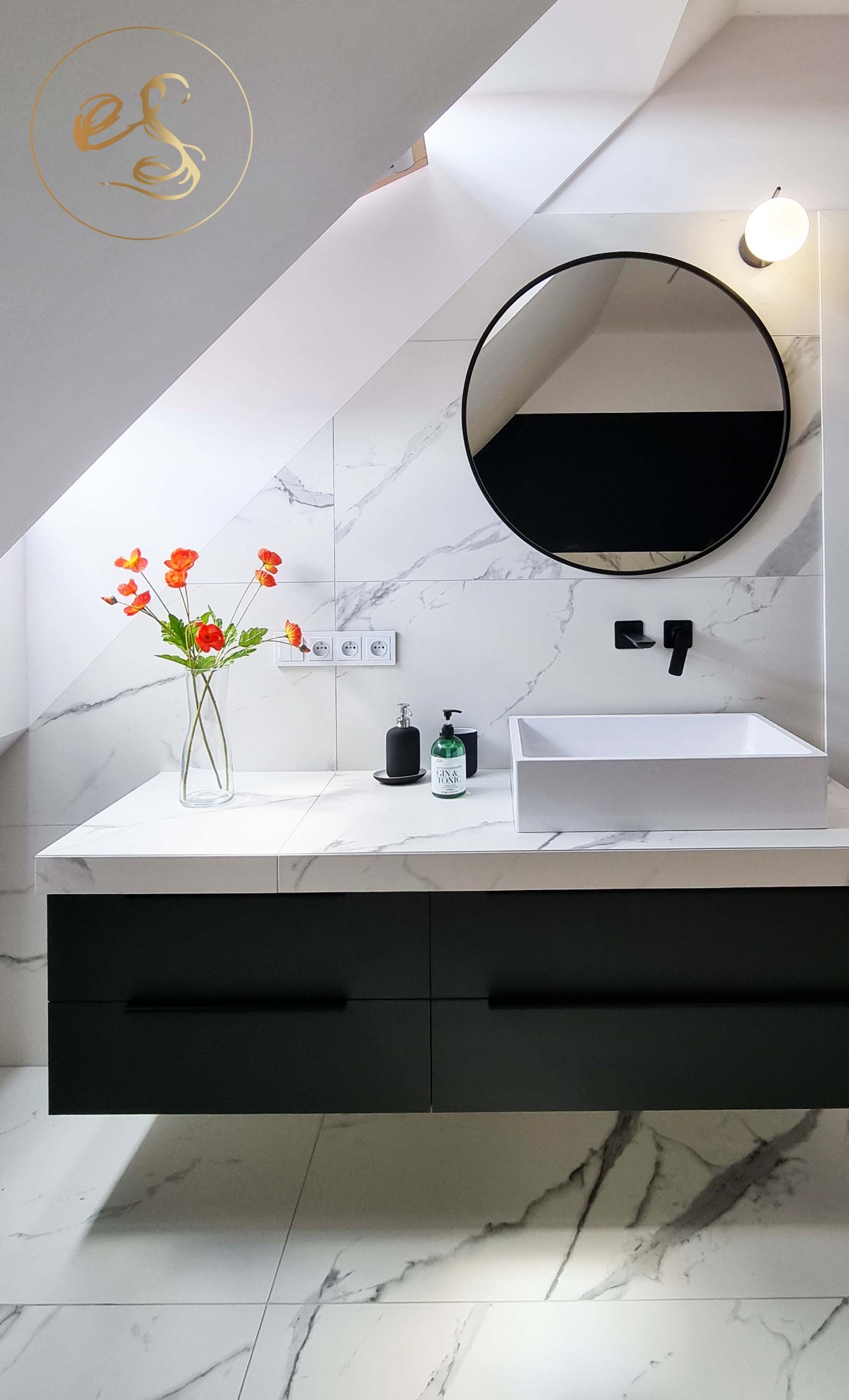 Nowoczesna łazienka na poddaszu z marmurowymi płytkami, okrągłym lustrem w czarnej ramie, białą umywalką nablatową i czarnymi szafkami pod umywalką, akcentem kolorystycznym jest bukiet...