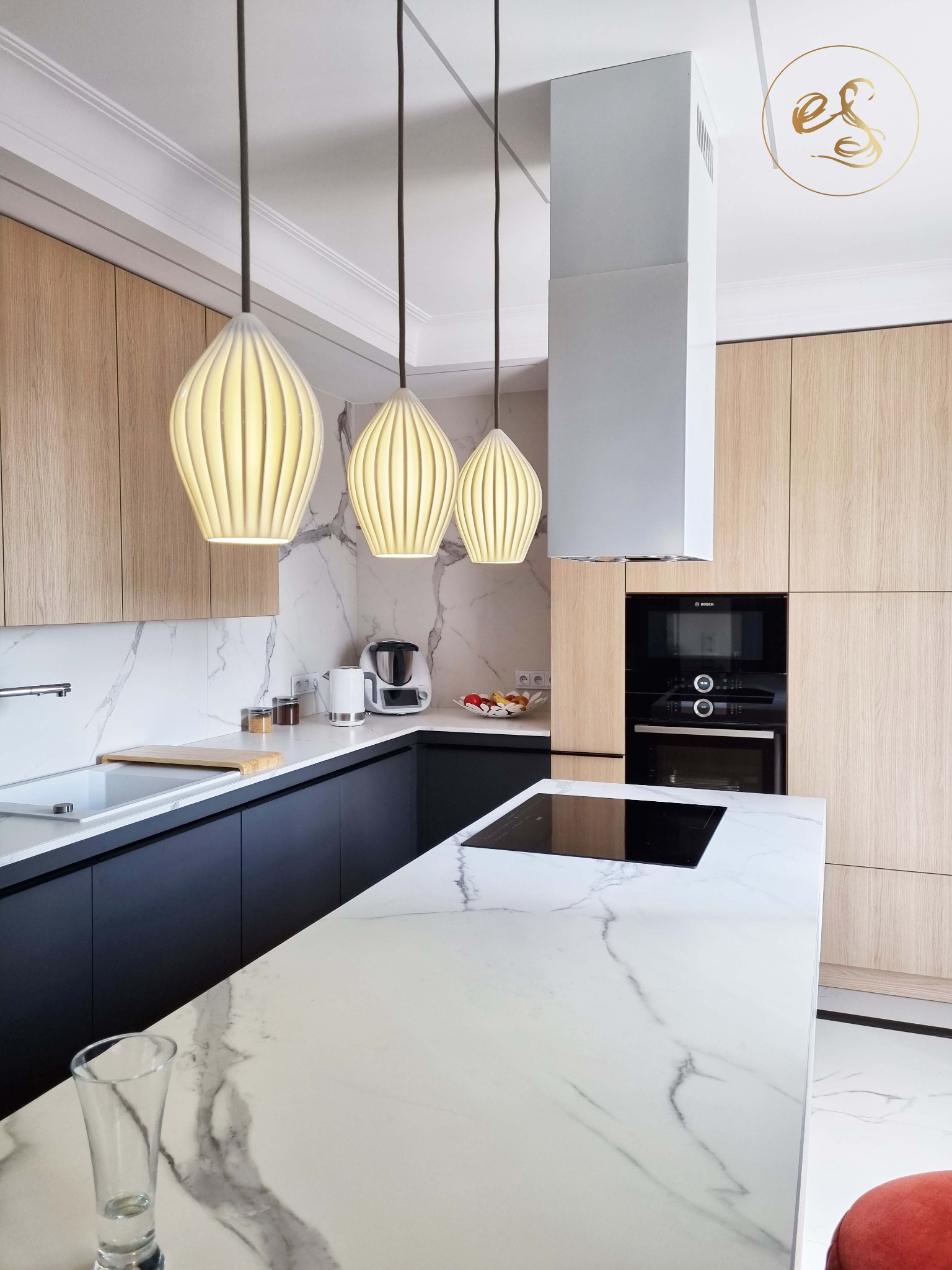 Nowoczesna kuchnia z marmurowym blatem wyspowym, drewnianymi szafkami i trzema wiszącymi lampami o organicznym kształcie, oświetlona naturalnym światłem.