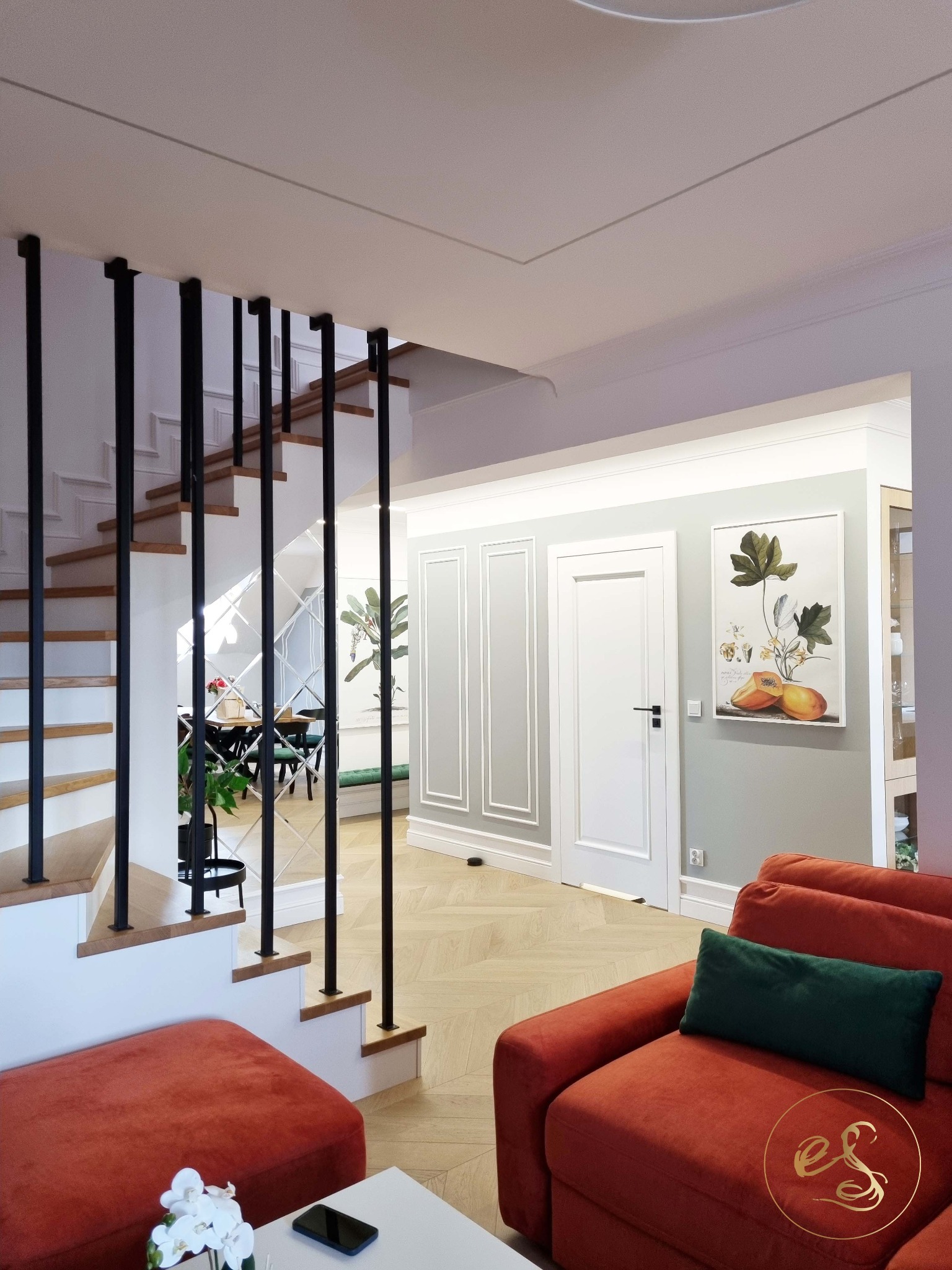 Eleganckie wnętrze domu z drewnianymi schodami z czarną balustradą, parkietem w jodełkę, pomarańczową sofą z zieloną poduszką, białym stołem z kwiatami i widoczną jadalnią w tle.