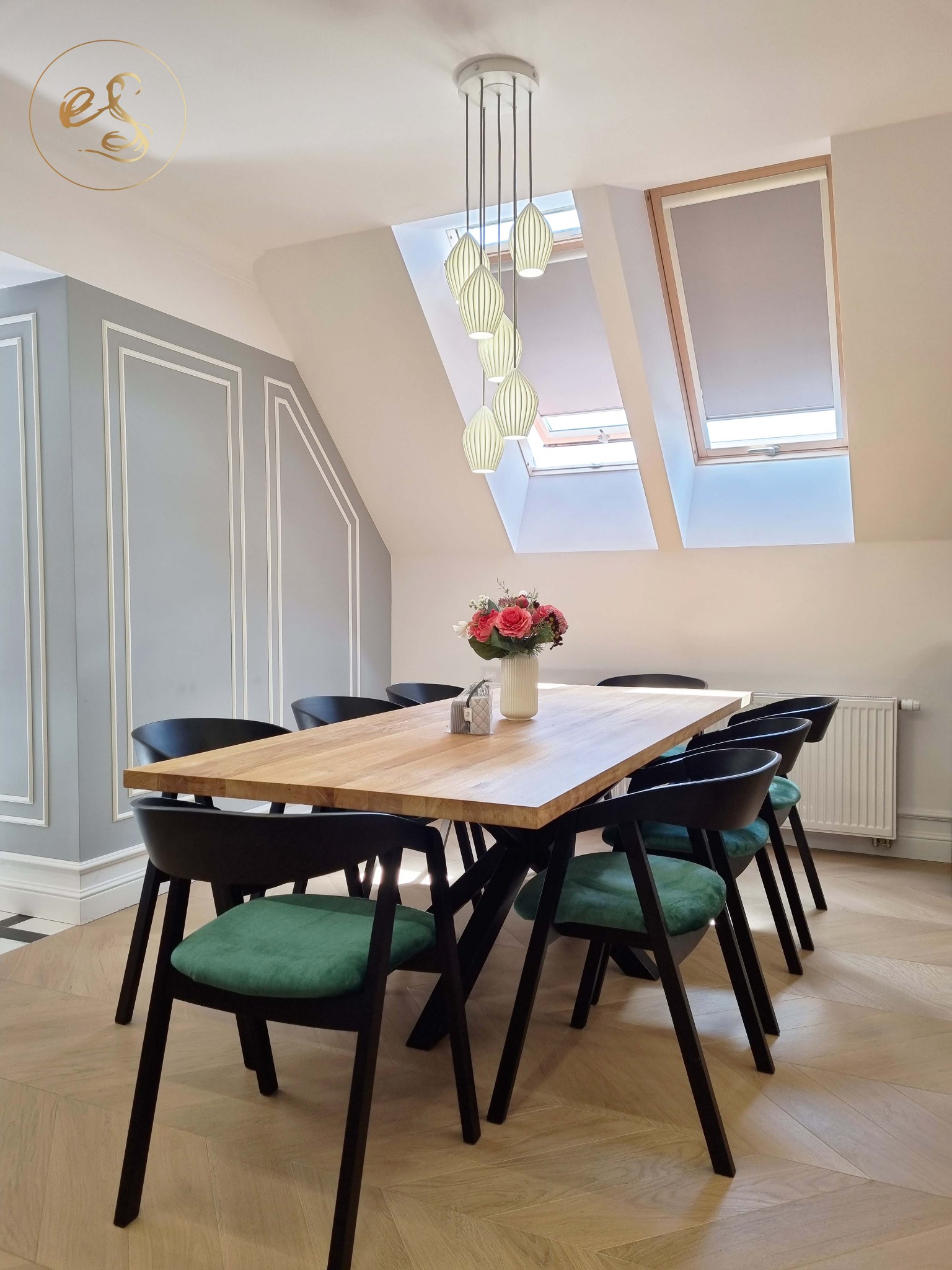 Jasna jadalnia na poddaszu z drewnianym stołem, czarnymi krzesłami z zielonymi siedziskami, bukietem róż w białym wazonie i wiszącą lampą z kilkoma kloszami