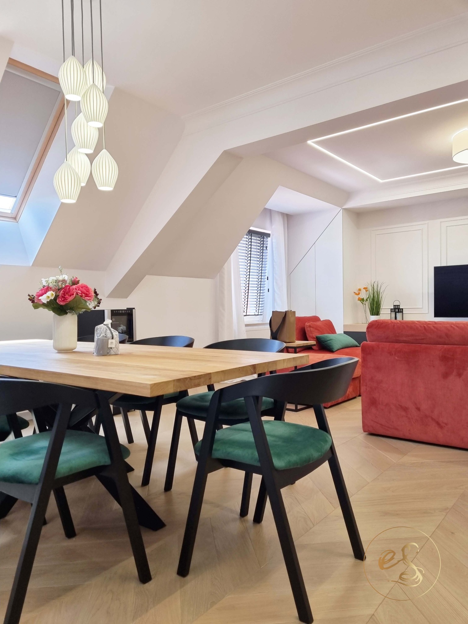 Jasne, przestronne poddasze z drewnianym stołem, zielonymi krzesłami i koralową sofą, oświetlone lampami wiszącymi i listwą LED w suficie.