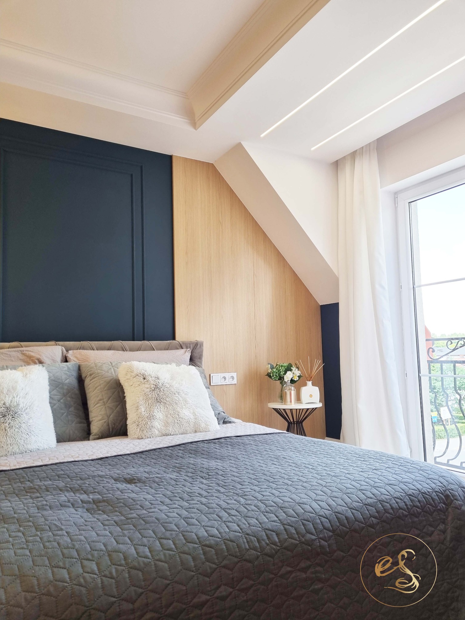 Sypialnia w odcieniach szarości i beżu z pikowaną narzutą, dekoracyjnymi poduszkami i kwiatami na stoliku nocnym przy balkonie.