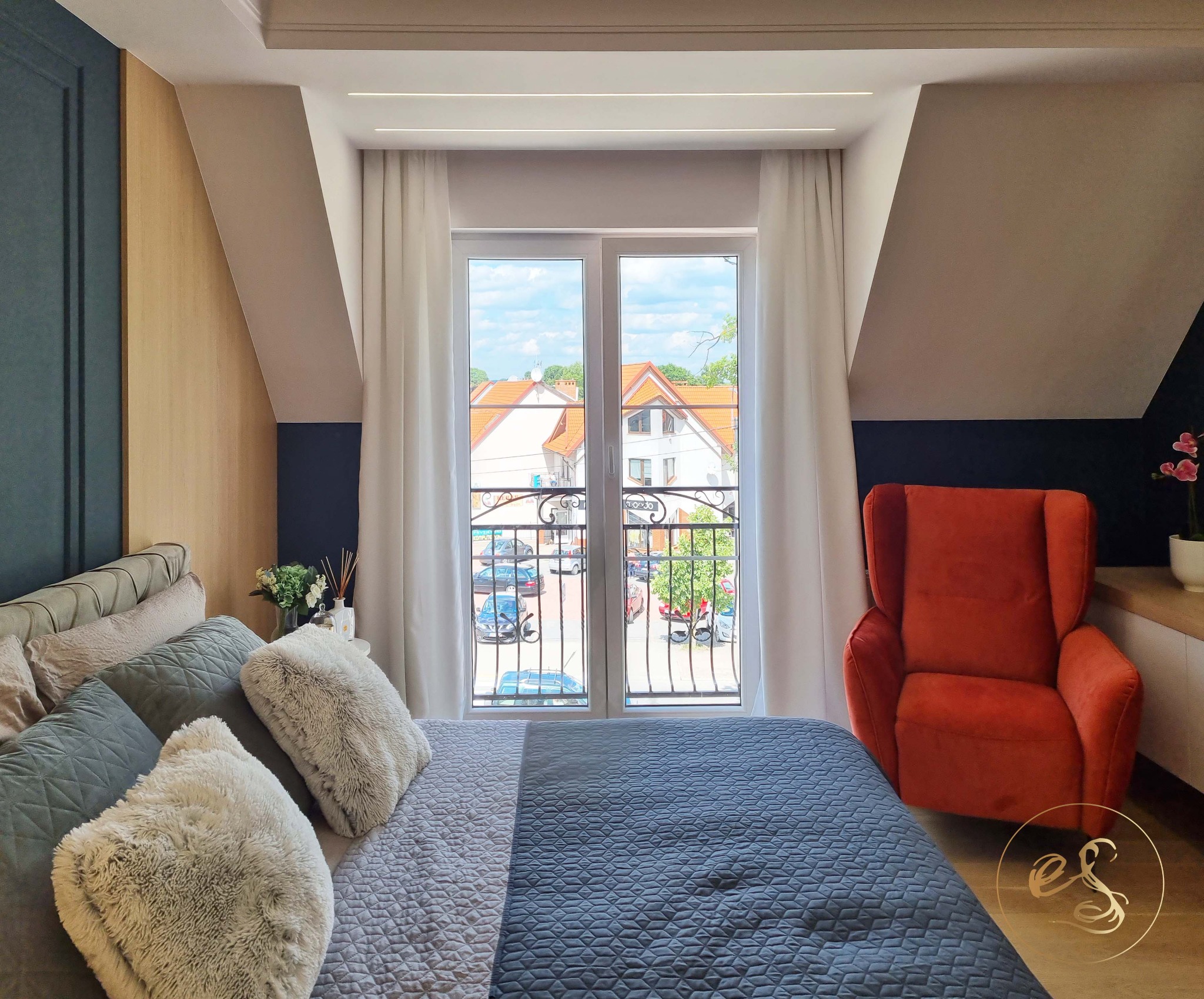 Jasna sypialnia na poddaszu z łóżkiem, pomarańczowym fotelem i widokiem na ulicę przez balkonowe okno z ozdobną balustradą.