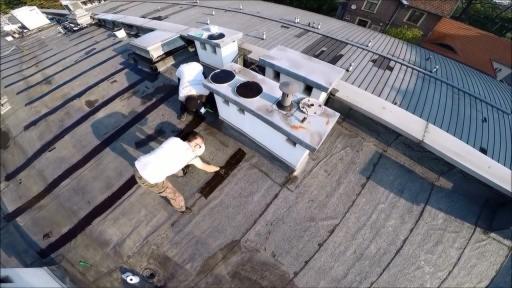 Dwóch pracowników uszczelnia płaski dach bitumiczny wokół kominów wentylacyjnych, widok z góry z szerokokątnym obiektywem.